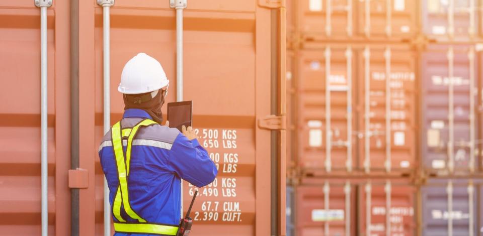 Port docker wearing hard hat and safety vest, standing in front of freight containers looking at supply chain trends on tablet