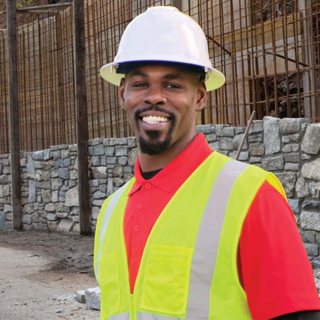 A man wearing a safety vest and hard hat standing in front of a stone wall in construction. 