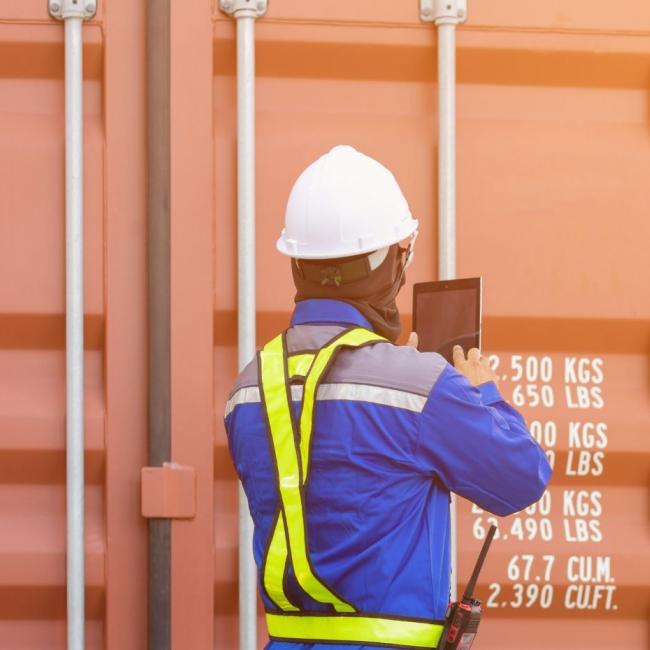 Port docker wearing hard hat and safety vest, standing in front of freight containers looking at supply chain trends on tablet